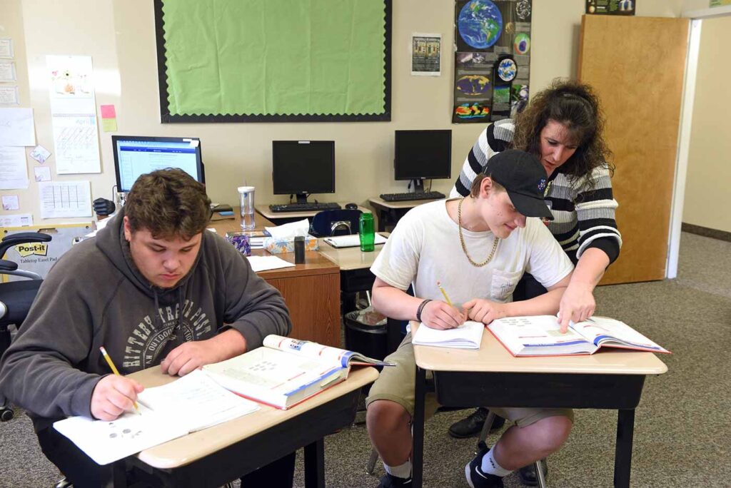Two teenage Natchaug students learn from a textbook while a teacher points out detail in textbook